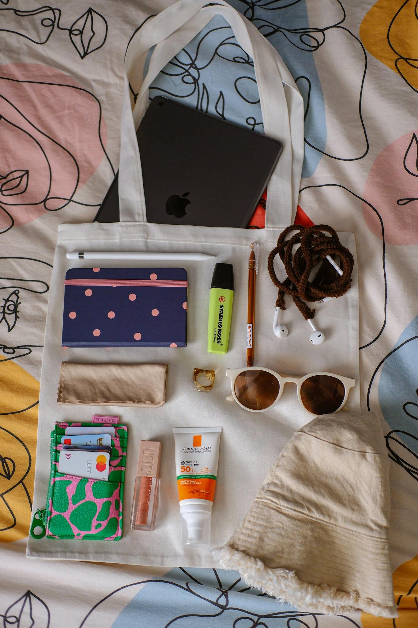 Flat lay of stylish tote bag with sunglasses, sunscreen, gadgets, and fashion accessories.
